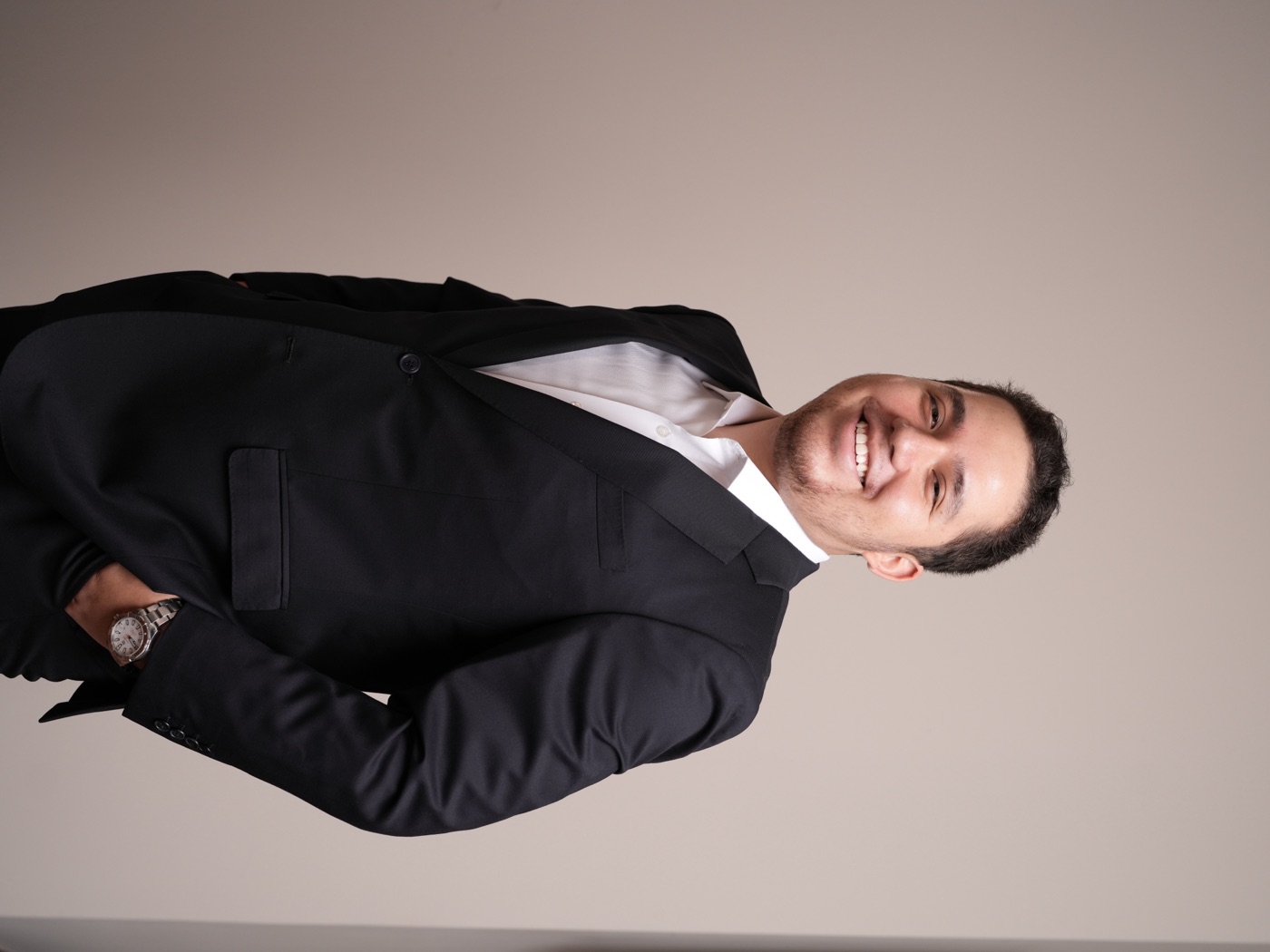 Portrait of Esteban Aranda smiling, wearing a dark suit and white shirt against a warm beige wall
